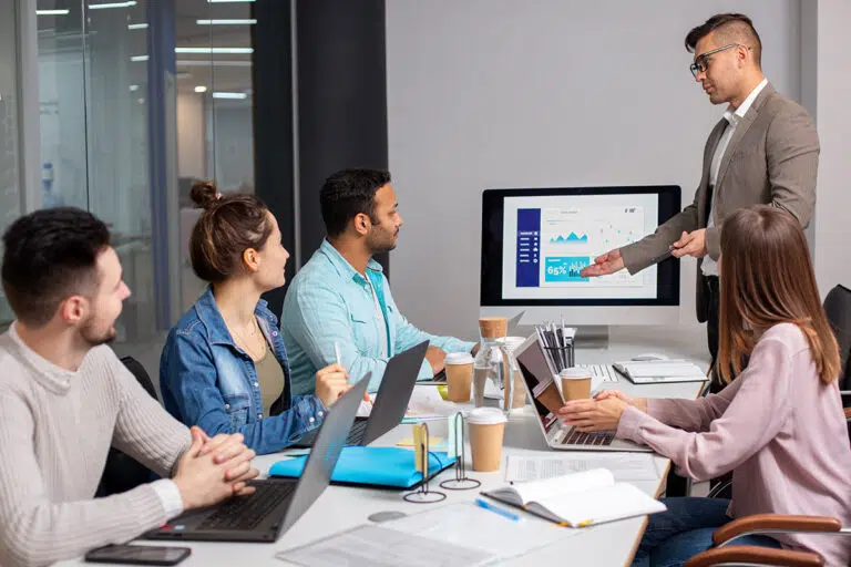 A man presents a bar chart on a monitor to four colleagues seated around a meeting table with laptops